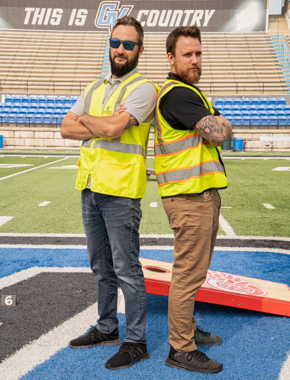 Jake Mallekoote and Matt DeRuiter standing back to back with their arms crossed, wearing construction vests and looking seriously into the camera